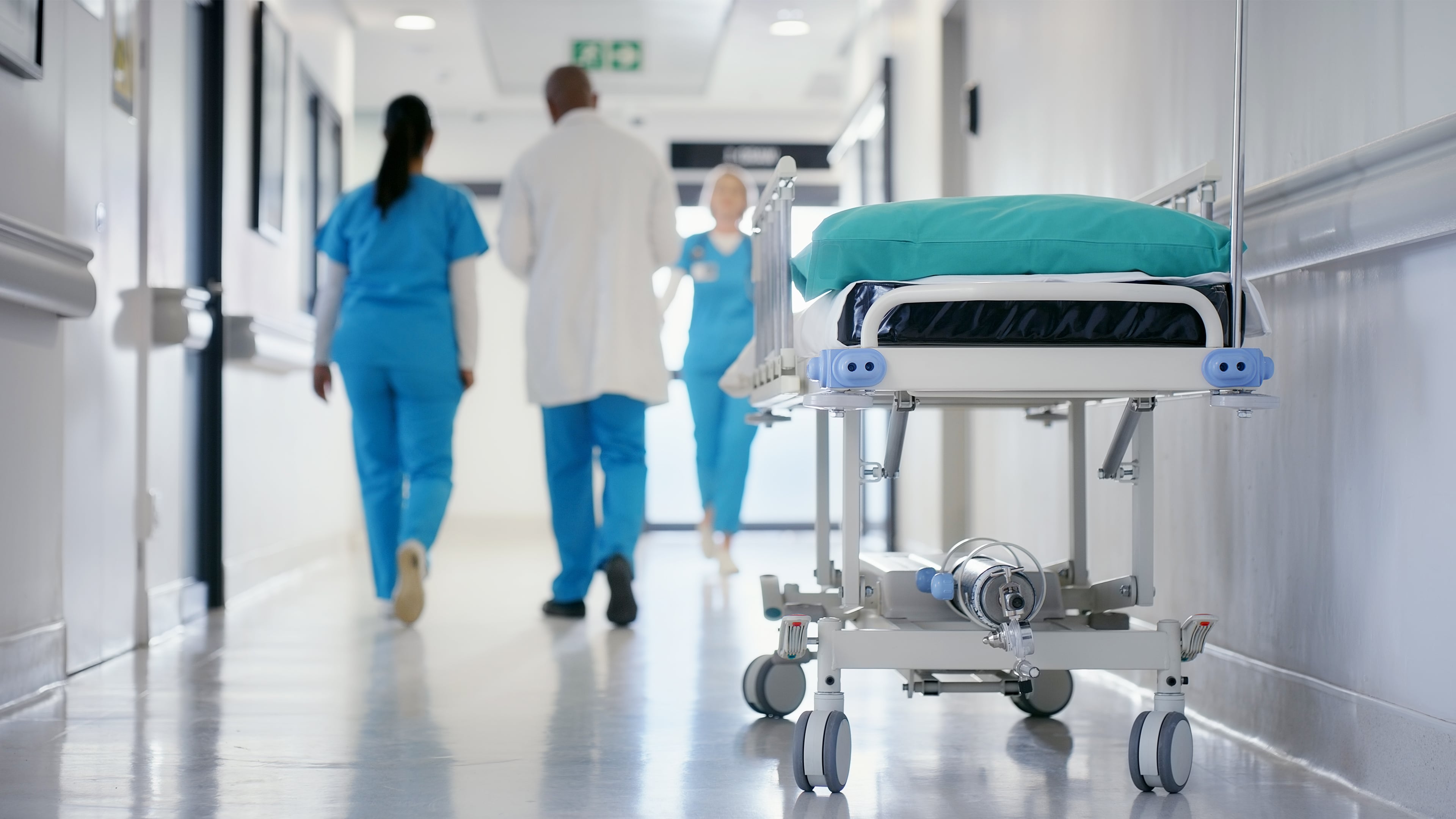 Real-life scenario image showing a hospital floor with three persons walking in the background and a hospital bed in front.