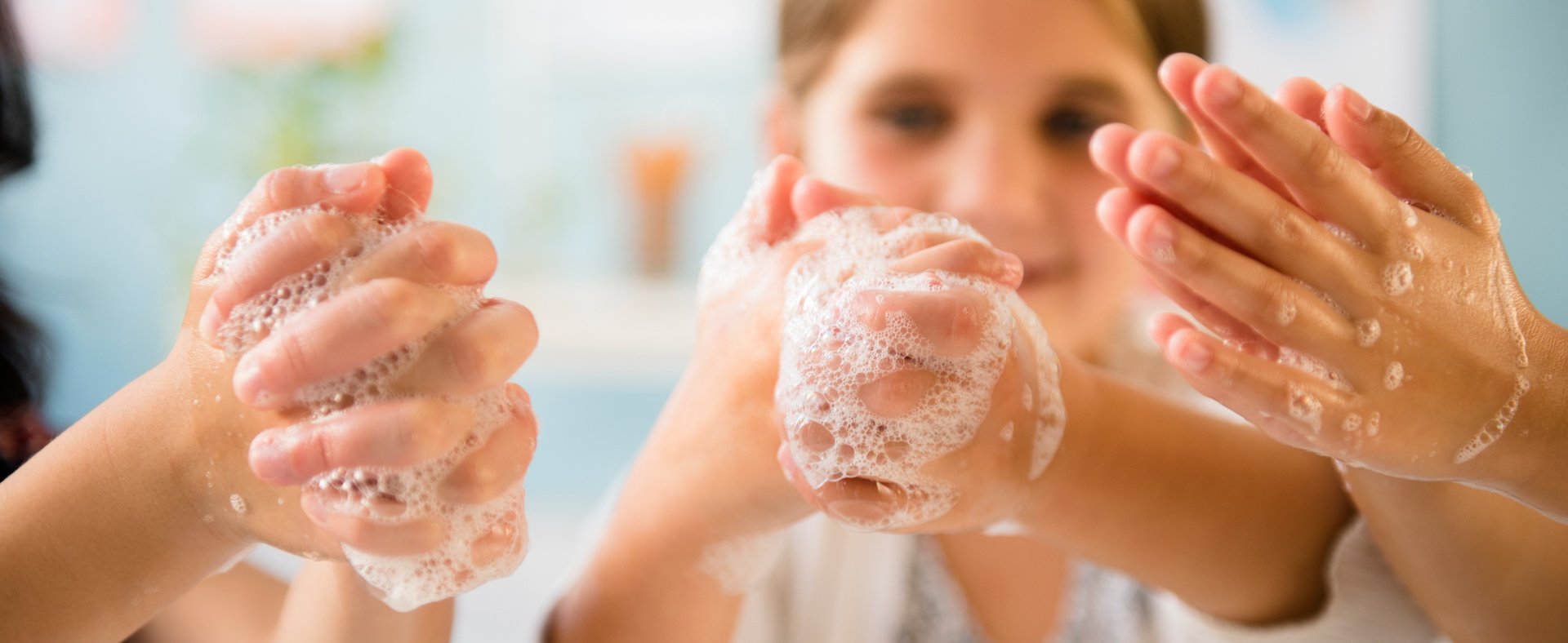 Three pair of hands with soap while washing the hands, a girl is seen in the background.