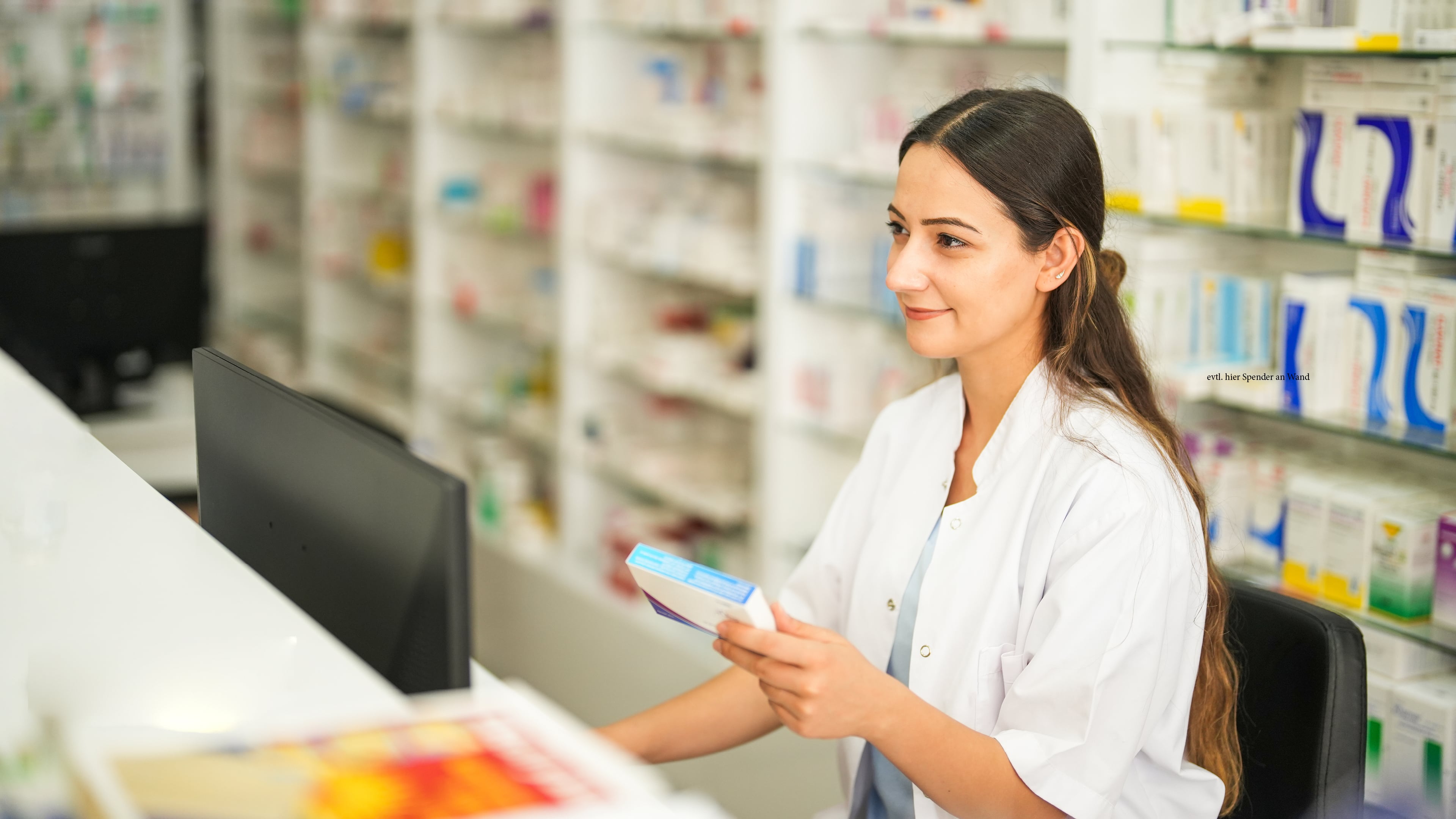 Pharmacy staff member holding a Hartmann medical product box while standing behind a counter with shelves of healthcare products in the background.