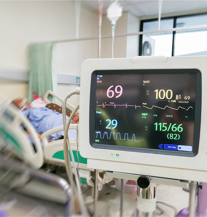 Patient monitoring device displaying vital signs beside a hospital bed in a clinical care setting.